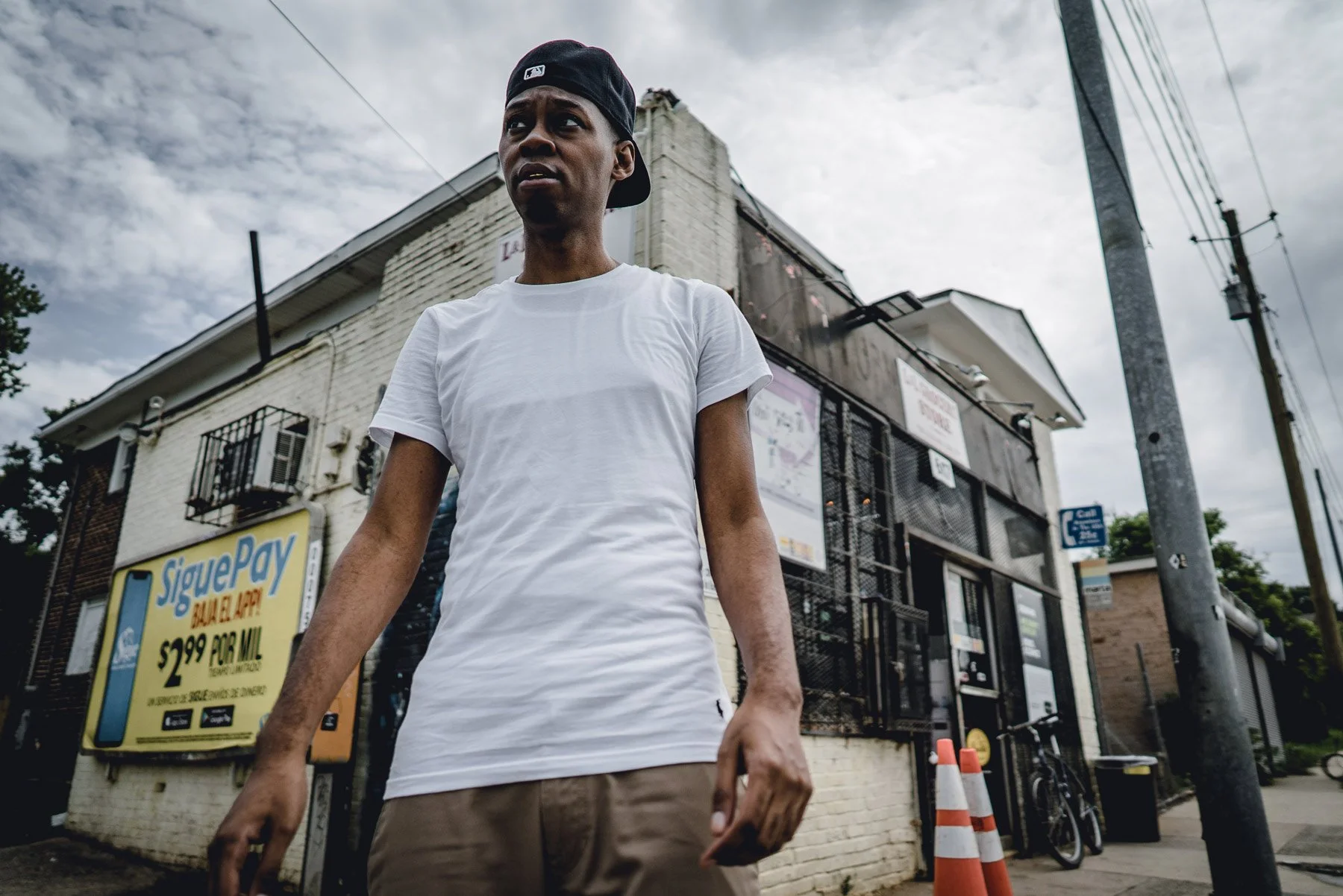 Cuz Lightyear wearing a white T-shirt and black cap on a city sidewalk