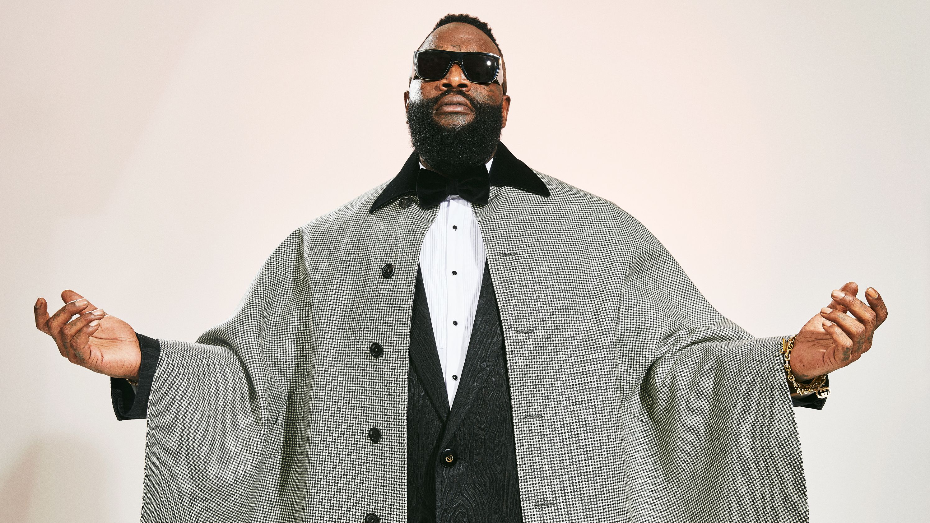 Rick Ross wearing sunglasses and a houndstooth cape in a studio portrait